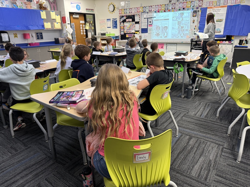 Elementary students sit at tables watching a classroom presentation projected on the board during a Junior Achievement lesson.