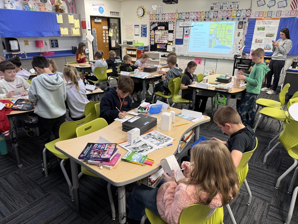 Students work independently at classroom tables, coloring and assembling paper buildings for a city-building activity.