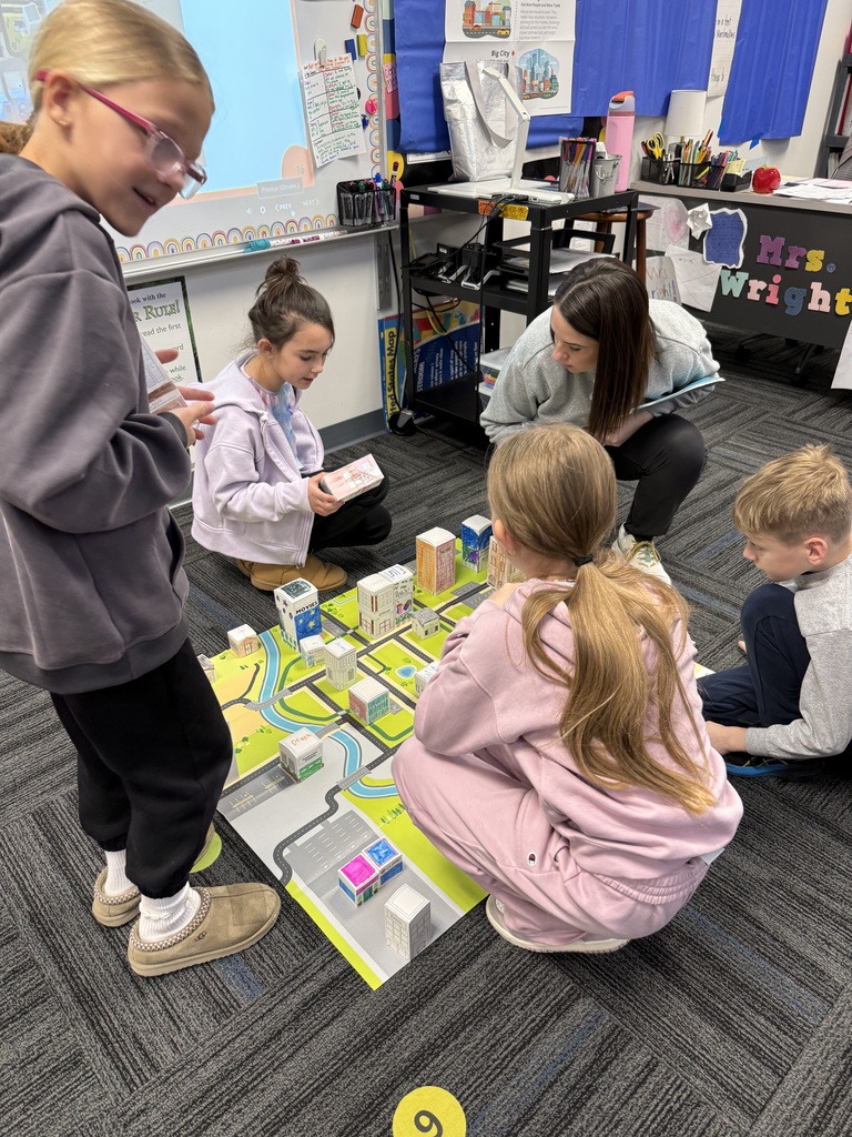 Small group of students and teacher collaborate around a city map, holding paper buildings for a city-planning activity.