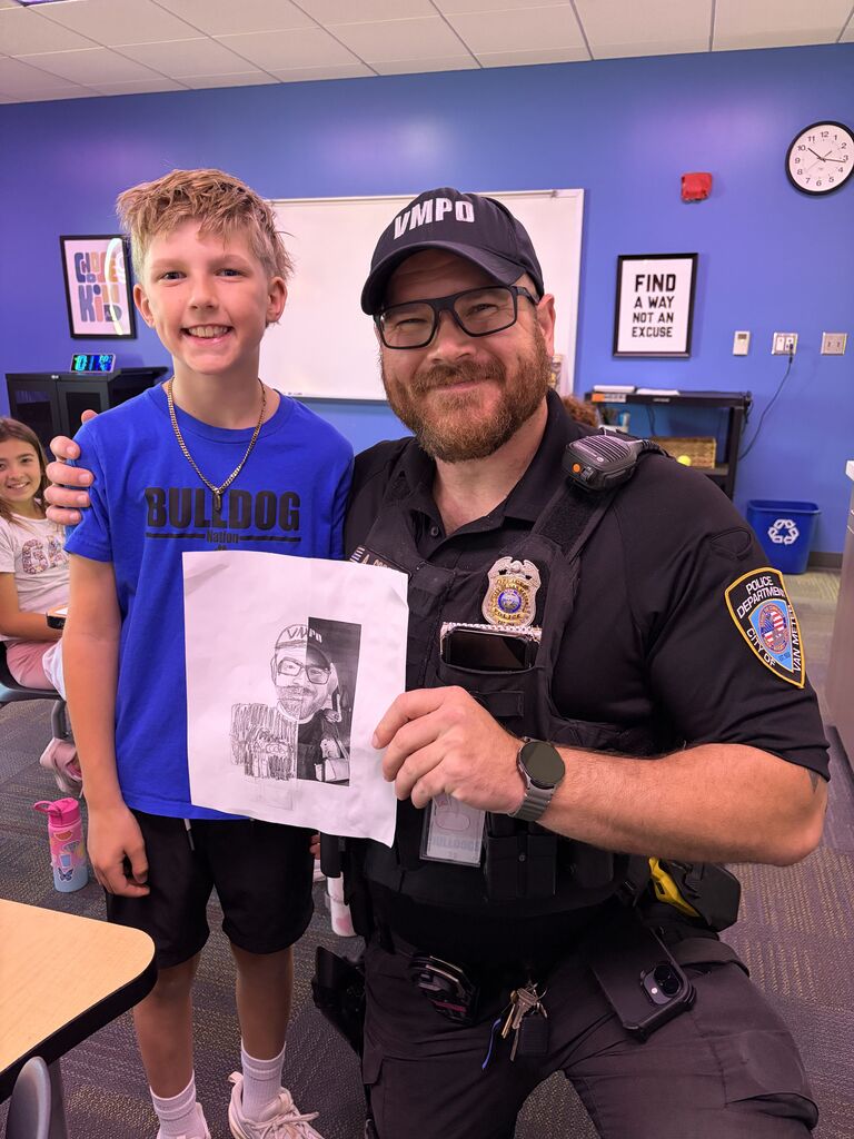 Smiling student and school police officer pose together holding a half-drawn portrait in classroom.