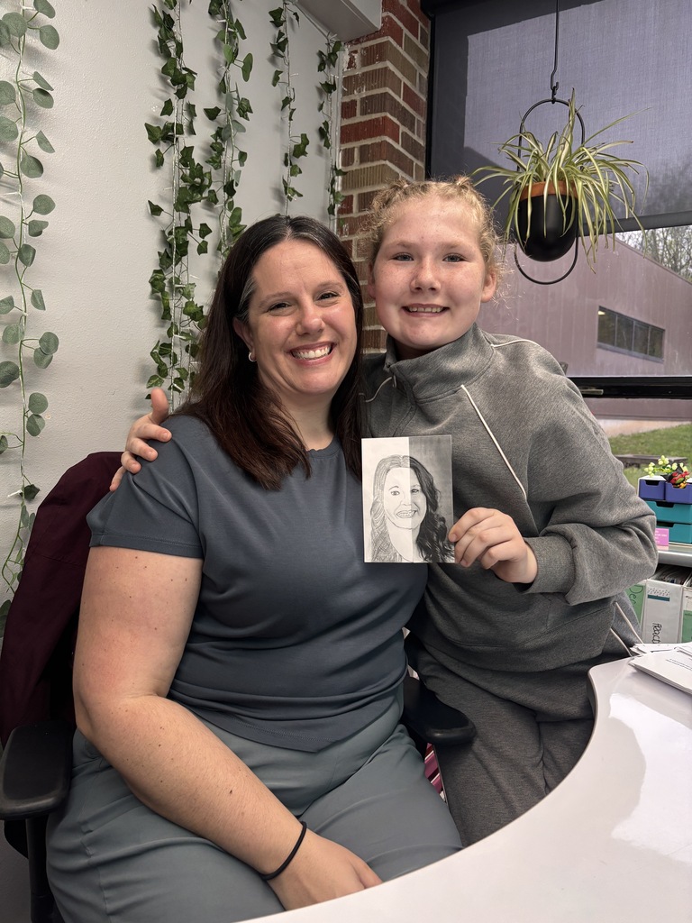 Student in gray hoodie stands beside smiling staff member holding a portrait drawing in office.