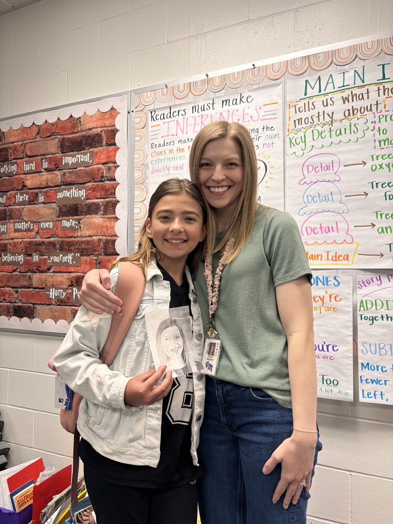 Student hugs smiling teacher while holding a portrait drawing in front of classroom posters.