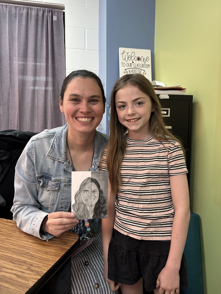 Young student and teacher smile while holding a completed half-drawing portrait in classroom.