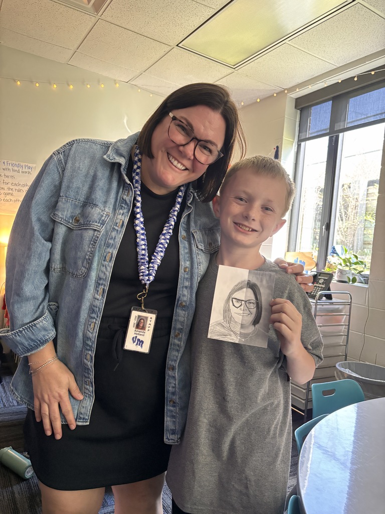 Student and teacher smile together while holding a half-drawn portrait sketch in a classroom.