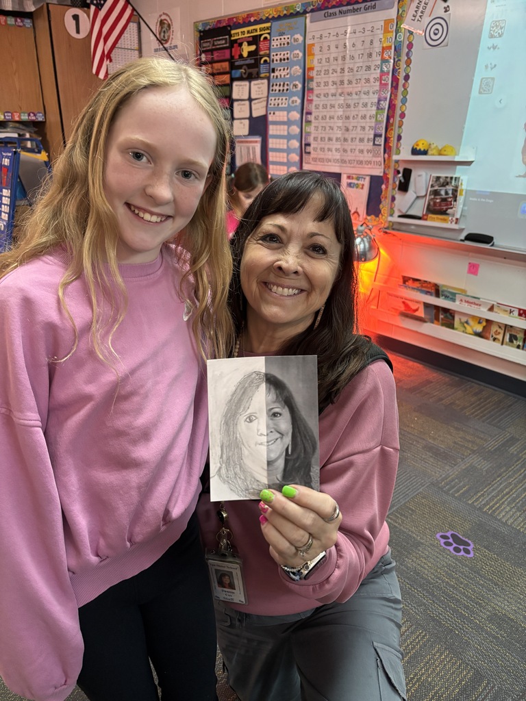 Student and teacher in pink tops smile while holding a completed half-drawn portrait artwork.
