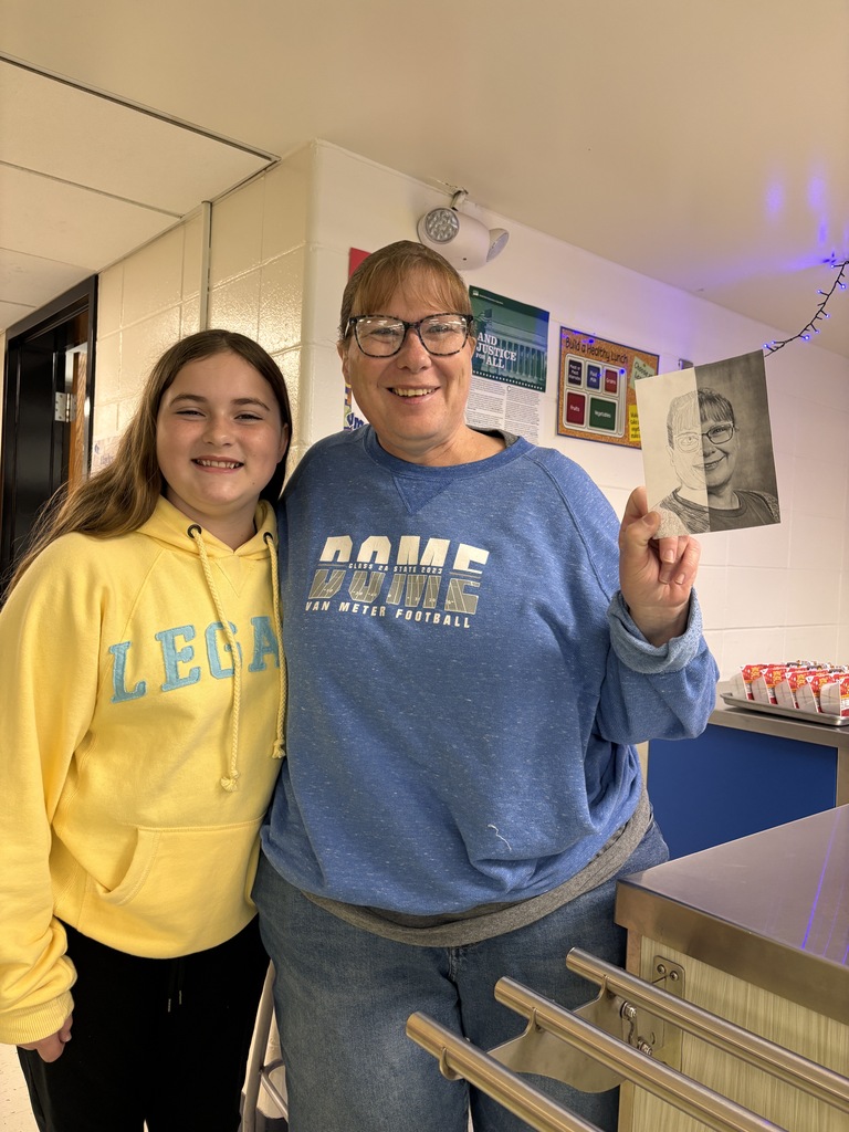 Student in yellow hoodie smiles beside staff member holding a finished portrait sketch in school cafeteria.