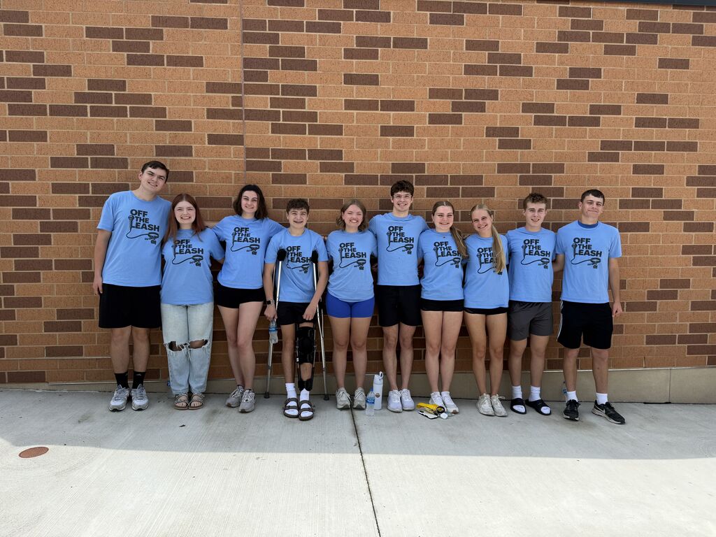 Group of ten teenagers standing side by side against a brick wall, all wearing matching light blue 'Off The Leash' T-shirts and casual bottoms.