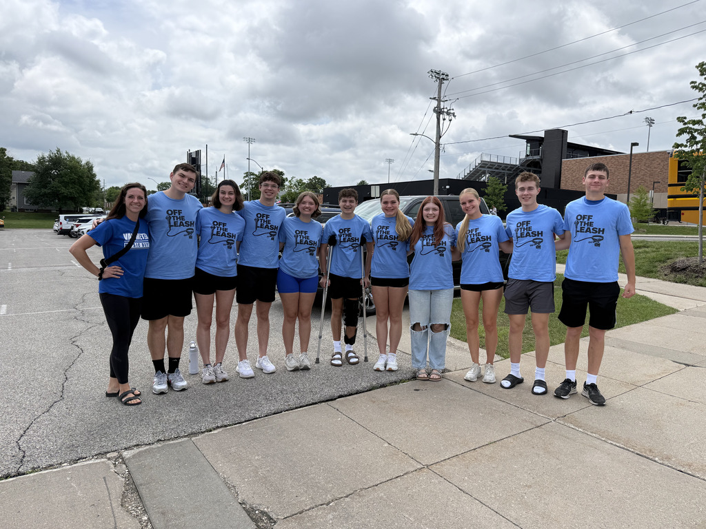 Group of eleven young people standing outdoors in a line, all wearing blue 'Off the Flash' t-shirts under a cloudy sky.