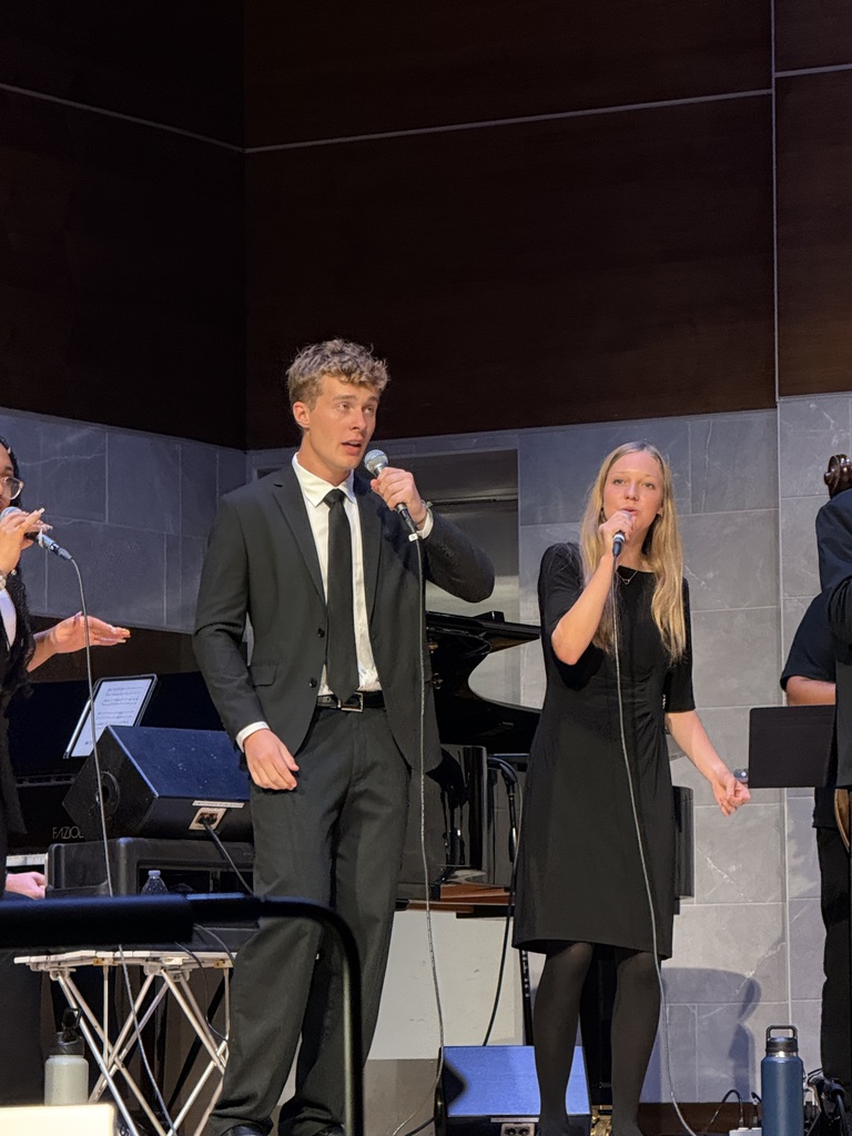 Two vocalists perform into handheld microphones onstage, dressed in black, with a grand piano behind them.