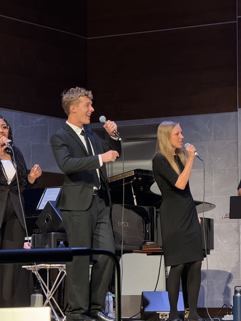 Two singers in formal attire performing on stage with microphones, accompanied by a pianist and a drum set in the background.