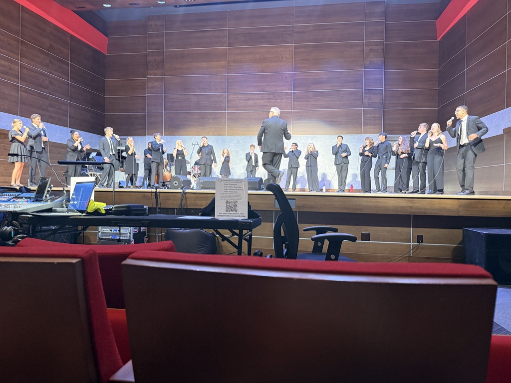 A vocal ensemble performs across a concert hall stage as a conductor leads from center, viewed from the audience behind the sound booth.
