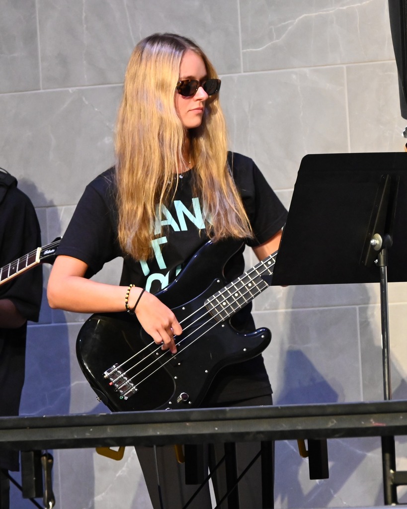 Person wearing sunglasses and a "Dance It Out!" shirt plays a black bass guitar beside a music stand during an indoor performance.