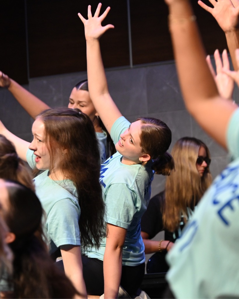 Show choir performers in matching "Dance It Out!" shirts raise their arms during a lively performance, while a bassist plays in the background.