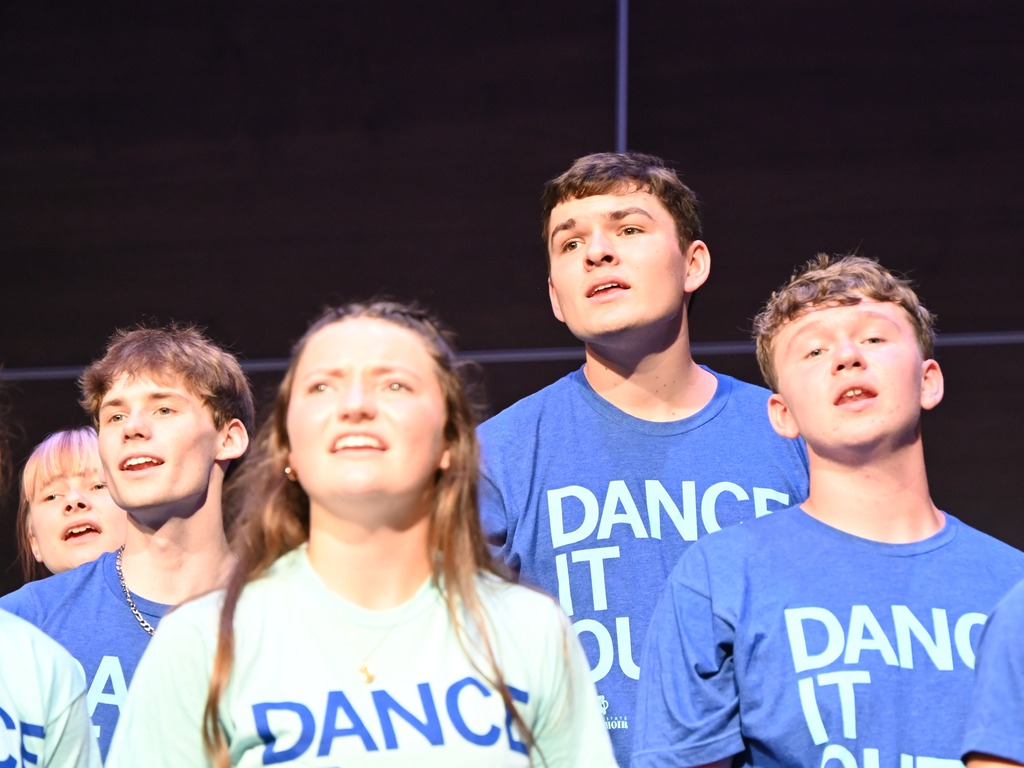 Show choir members in matching "Dance It Out!" shirts sing together onstage, looking upward during an expressive group performance.