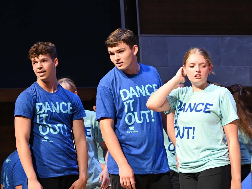 Three show choir performers in matching "Dance It Out!" shirts act out a scene onstage while fellow cast members stand in the background.