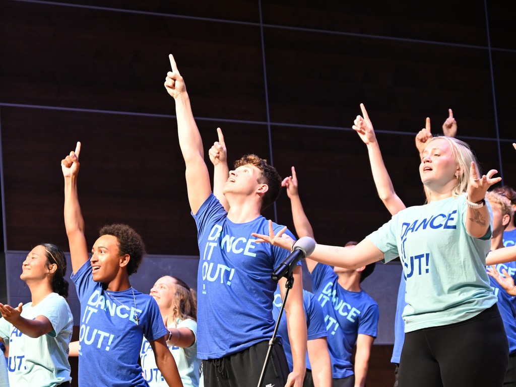 Performers in "Dance It Out!" shirts sing onstage with arms raised and fingers pointing upward during an energetic group performance.