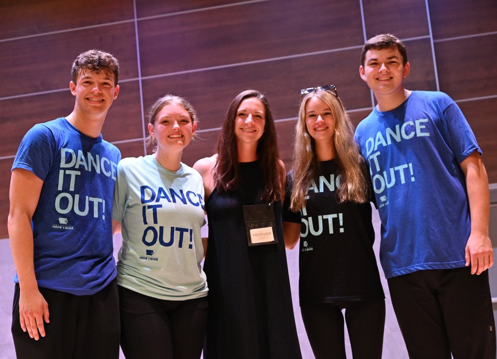 Five smiling people pose together indoors, with four wearing “Dance It Out!” show choir shirts beside an instructor wearing a name badge.