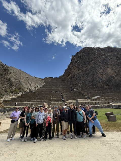 Group of tourists posing in front of ancient stone terraces carved into a mountainside under a partly cloudy sky.