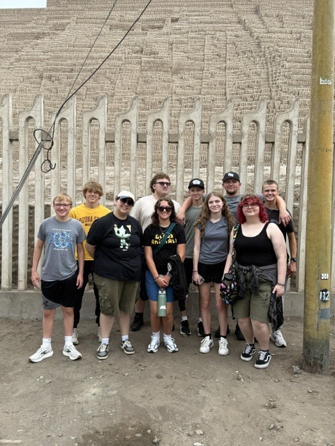 Group of ten young adults posing in front of an ancient stone wall with vertical carved columns outdoors.