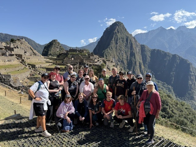 Group of travelers posing at Machu Picchu with the iconic mountain peak and sunlit Andes rising behind the ancient stone ruins.