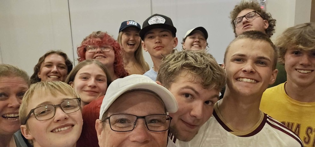 Group selfie of diverse young people and an older man smiling indoors against a plain wall.
