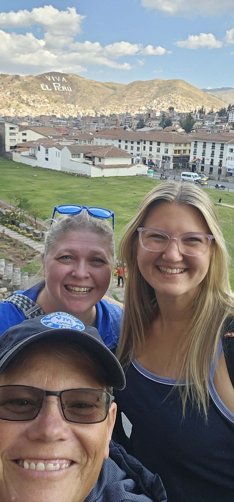 Three smiling people taking a selfie with a cityscape and mountains in the background under a partly cloudy sky.