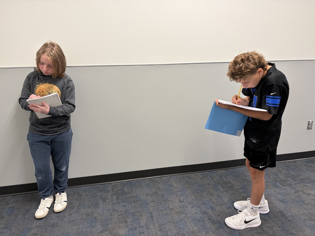 Two students stand apart in a hallway, looking down as they write in notebooks.