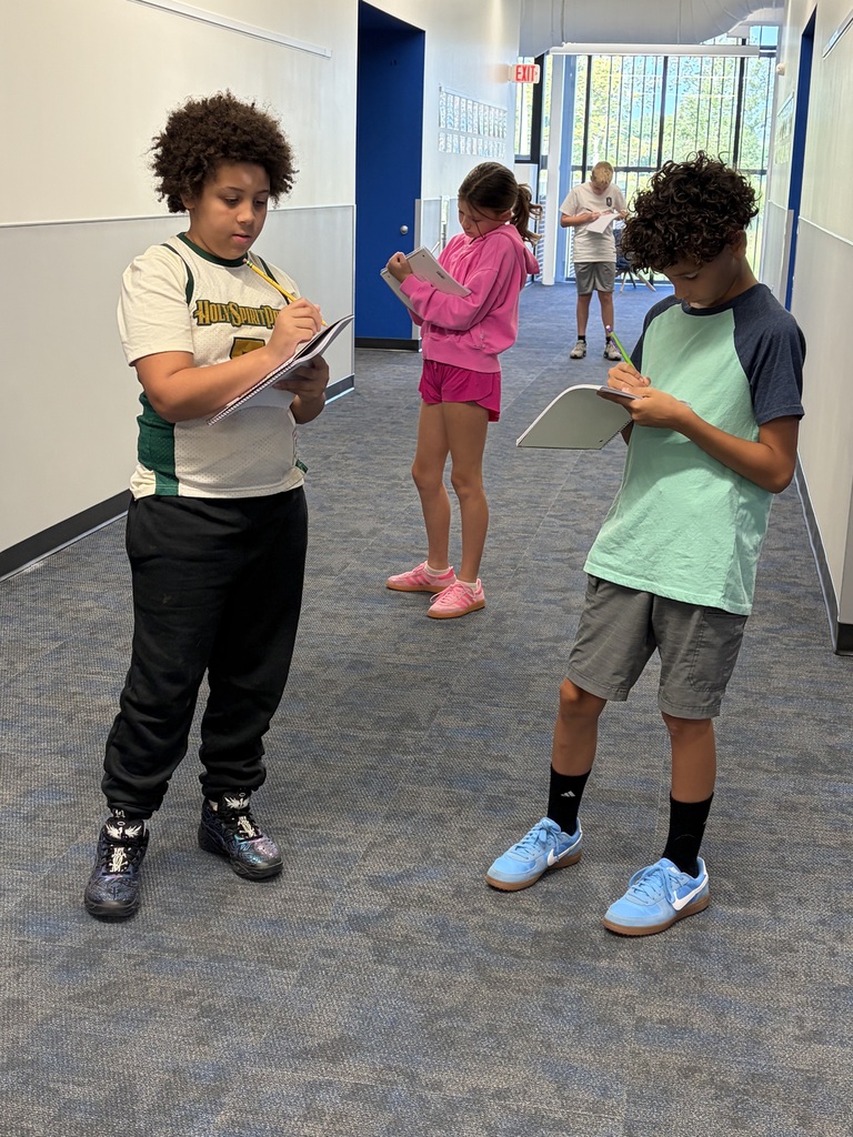 Three children standing in a hallway, each focused on writing or drawing on clipboards.