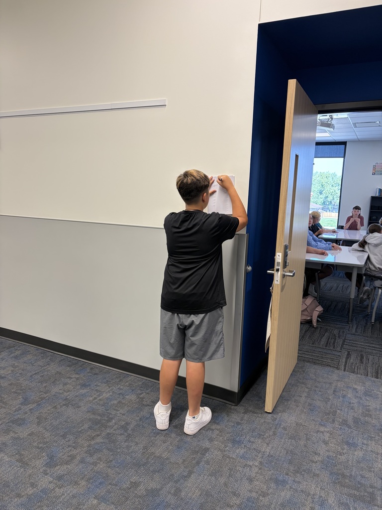 A boy in a black shirt and gray shorts writing on a paper taped to a white wall near an open door to a classroom.