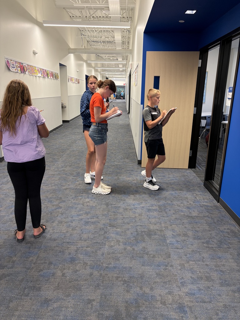 Four students standing in a school hallway, each focused on writing in notebooks while spaced apart.