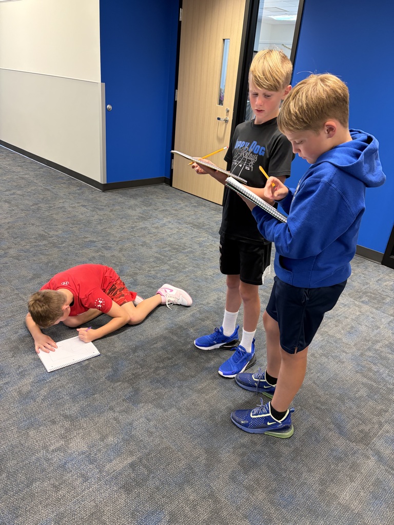 Three boys engaged in a school activity, two standing and writing in notebooks while one lies on the floor reading a paper.