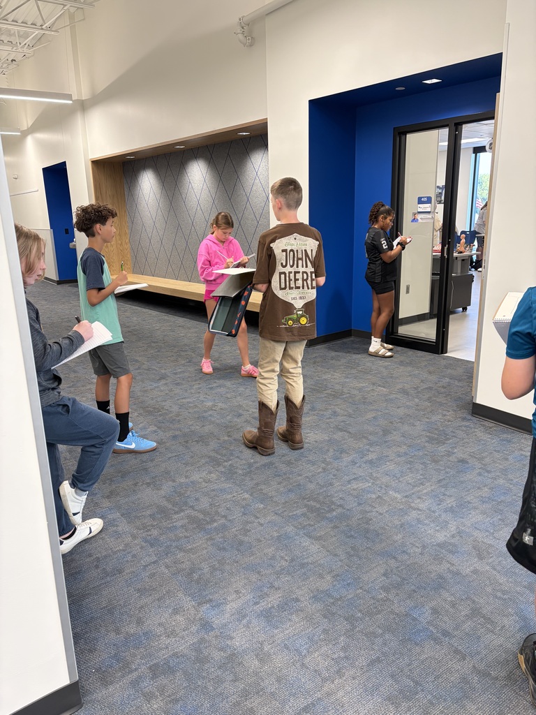 Children standing and writing on clipboards in a school hallway with blue and white walls and carpeted floor.