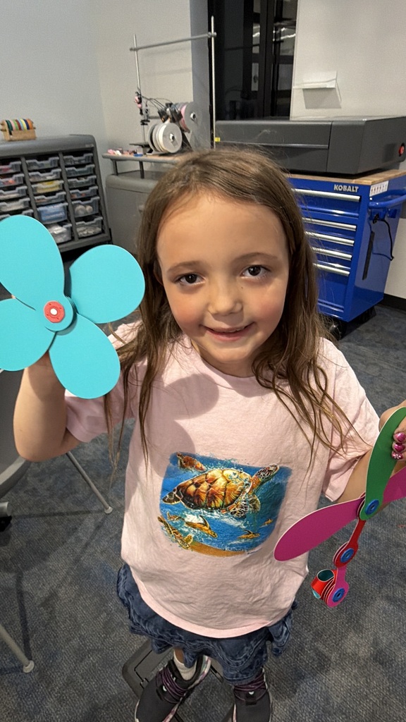 Smiling young girl holds two colorful handmade propeller crafts in a workshop filled with tools and supplies.