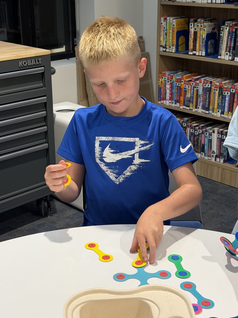 Young boy in a blue Nike shirt assembling colorful plastic pieces at a white table in a library setting.