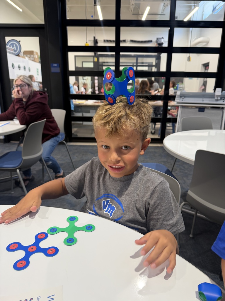Young boy with a colorful fidget spinner crown on his head, sitting at a table with more fidget spinners in front of him in a classroom setting.