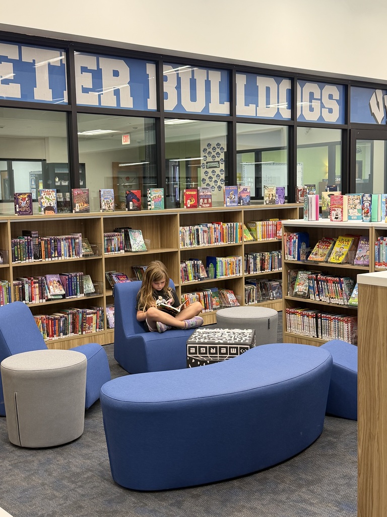 A young girl reading a book while sitting on a blue chair in a school library filled with bookshelves and colorful book displays.