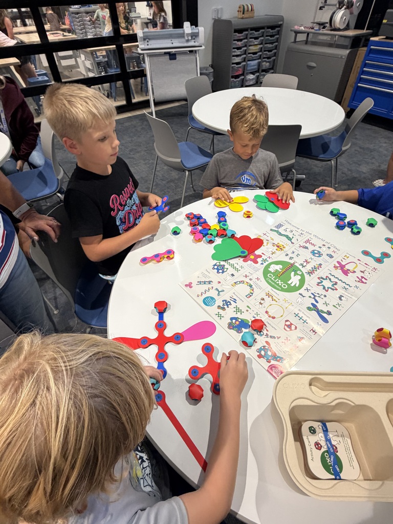 Three young boys engaged in a colorful educational activity with shapes and tokens on a round table in a classroom.