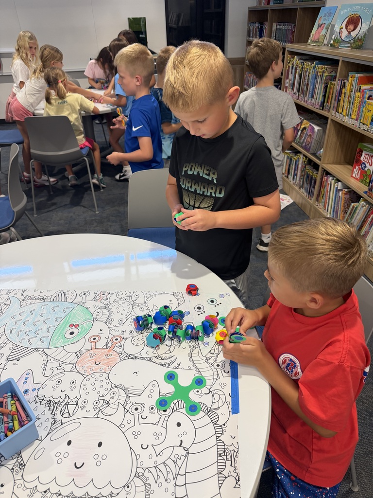 Two boys playing with colorful fidget spinners on a large sea creature coloring sheet in a library setting.