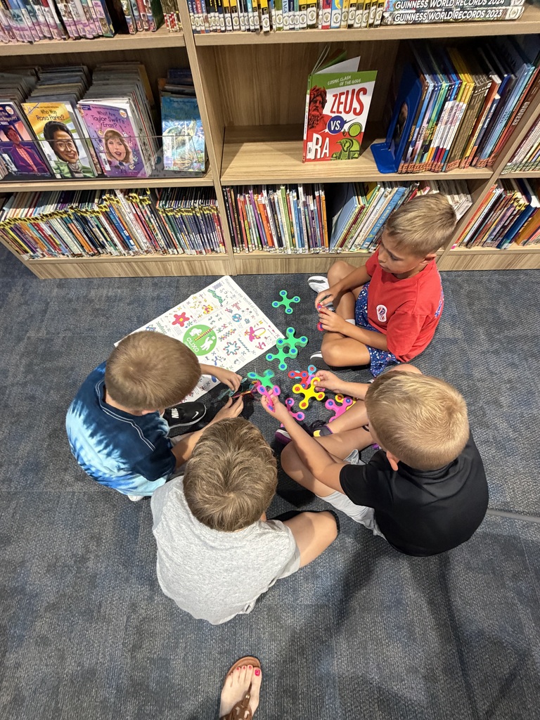 Four young boys sitting on a library carpet floor playing with colorful fidget spinners near a bookshelf filled with children's books.