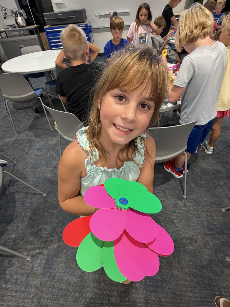 Smiling young girl holding colorful paper flower petals in a classroom with children crafting in the background.