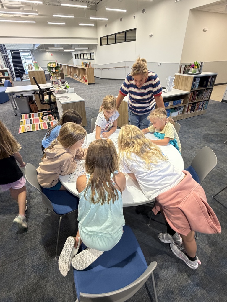 A group of young children gathered around a table in a modern classroom, engaged in a collaborative activity with a teacher guiding them.