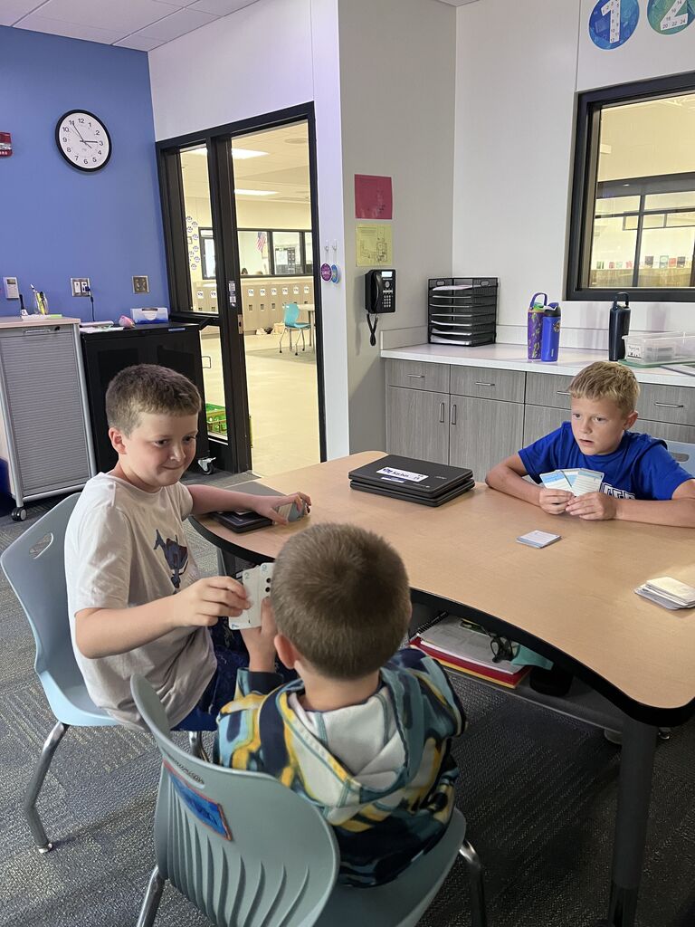 Three students sit around a classroom table holding number cards while playing an interactive math game.