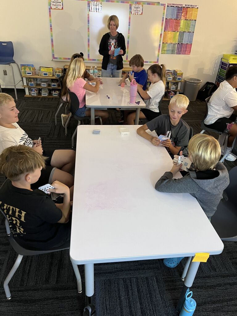 Students play a card-based math game at classroom tables while a teacher stands nearby holding a set of cards.