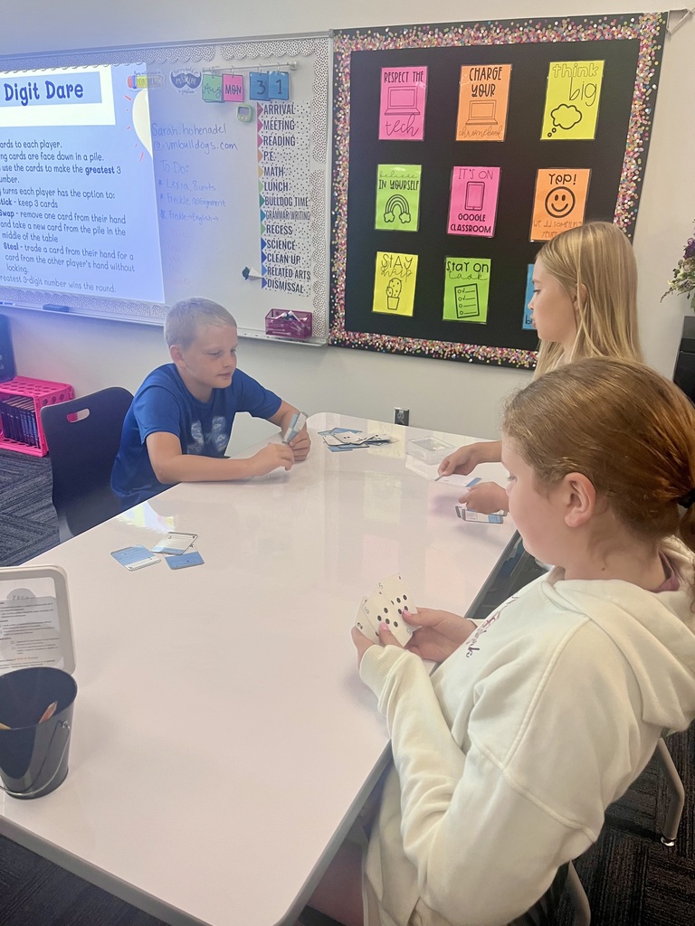 Three students use number cards at a classroom table while participating in the projected “Digit Dare” math activity.