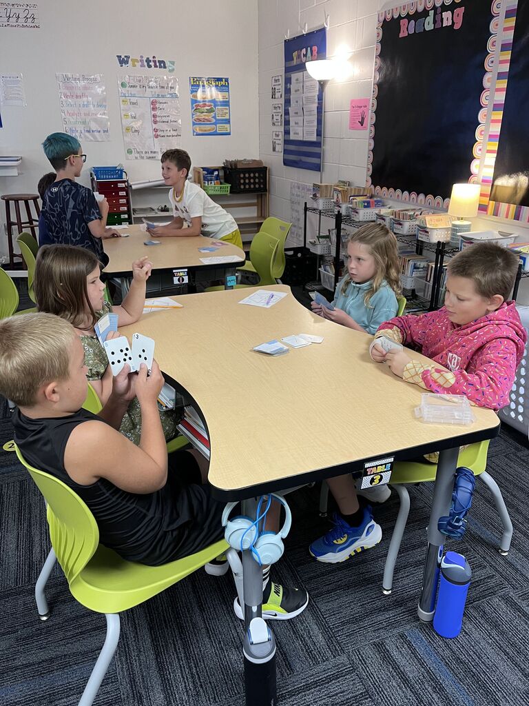 Students work in small groups at classroom tables, holding number cards and participating in a math game.