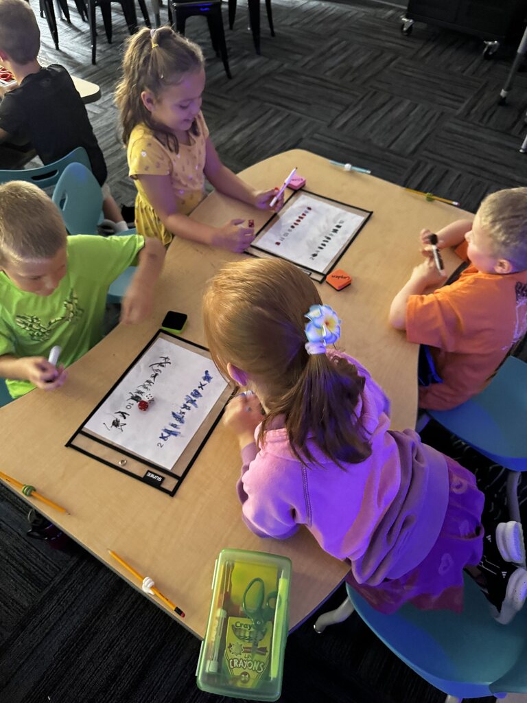 Four students gather around a table, using markers, dice, and dry-erase boards during a hands-on math activity.