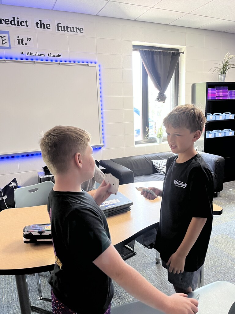 Two students stand across from each other in a classroom, comparing cards during a math activity.