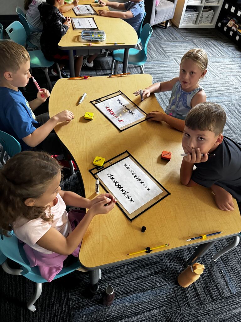 Four students gather around a classroom table, using dry-erase boards and markers to practice math together.