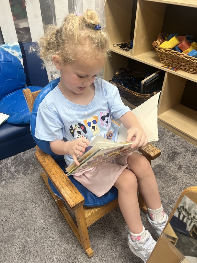 Young girl with curly blonde hair sitting in a wooden chair, focused on reading a book indoors.