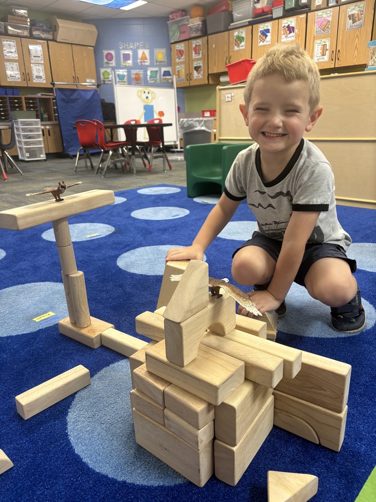 Young boy smiling while playing with wooden blocks in a colorful classroom with educational posters and furniture.
